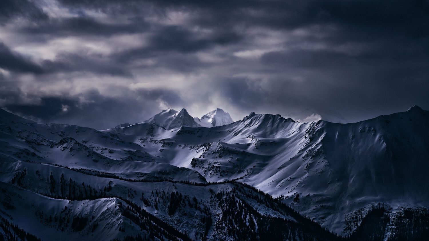 Dramatic snow-capped mountain peaks under dark stormy clouds in a moody winter landscape.