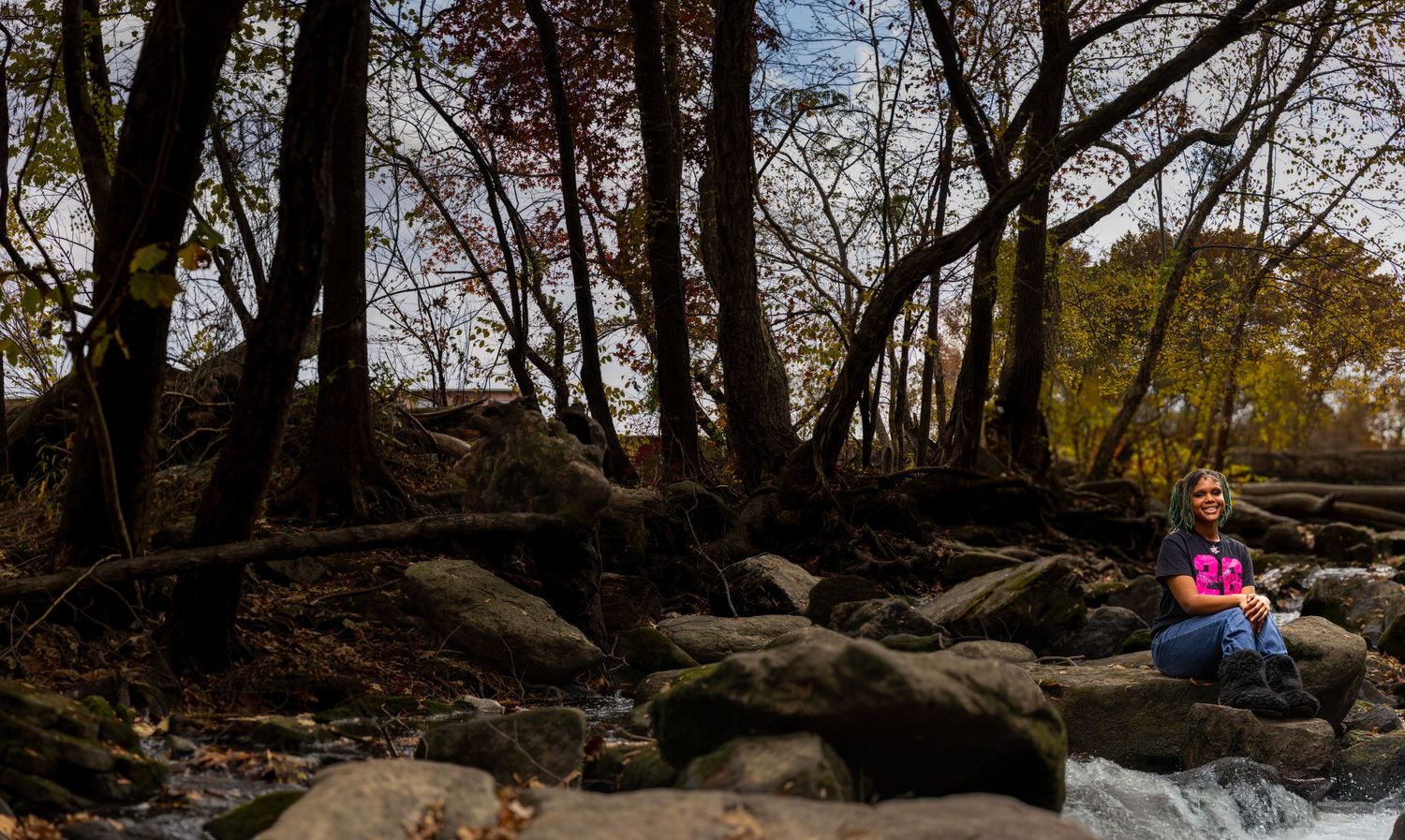 Person sitting on rocks beside a stream in an autumn forest setting.
