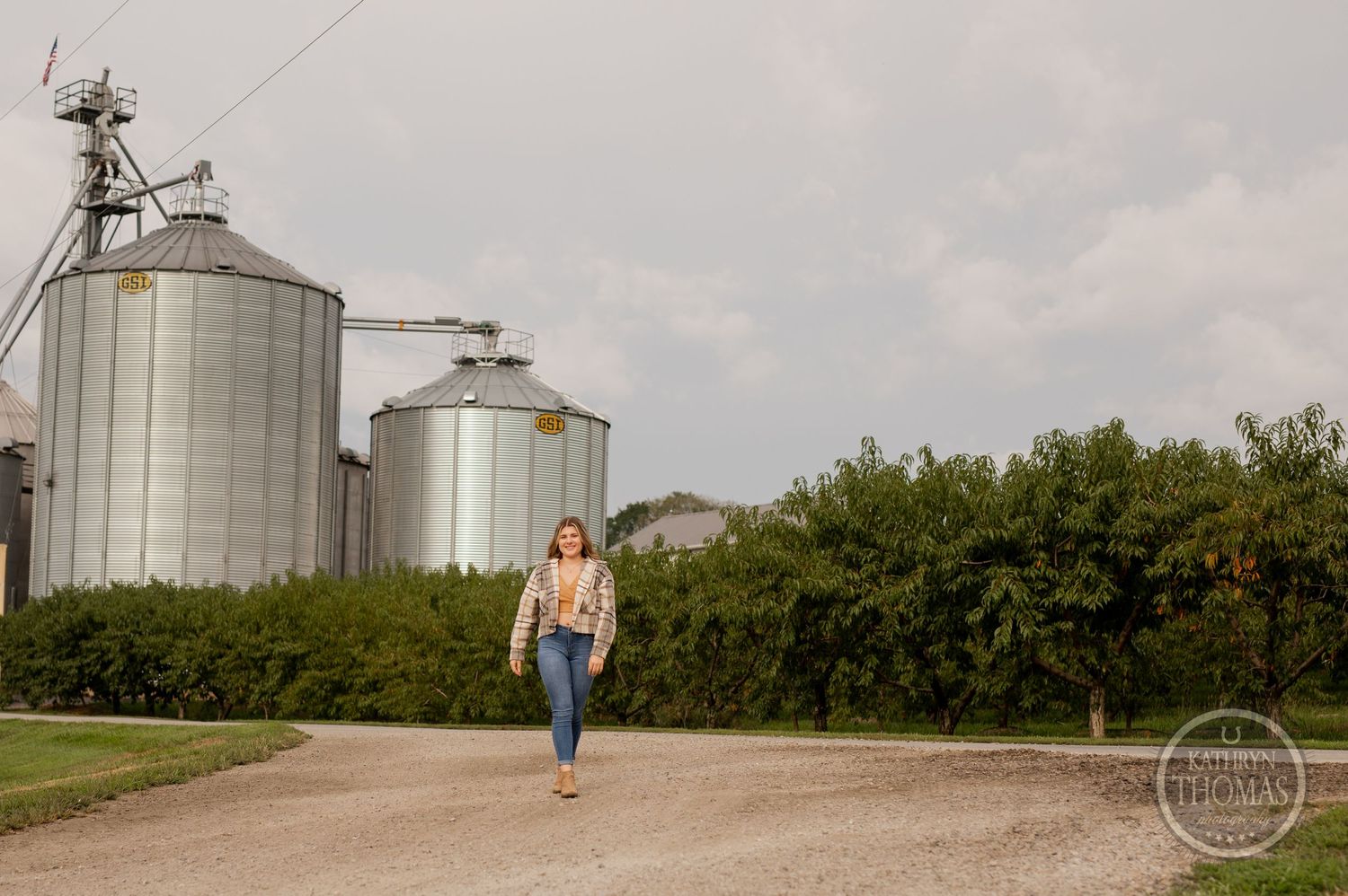 Person in denim walks on dirt path with large grain silos in background on cloudy day.