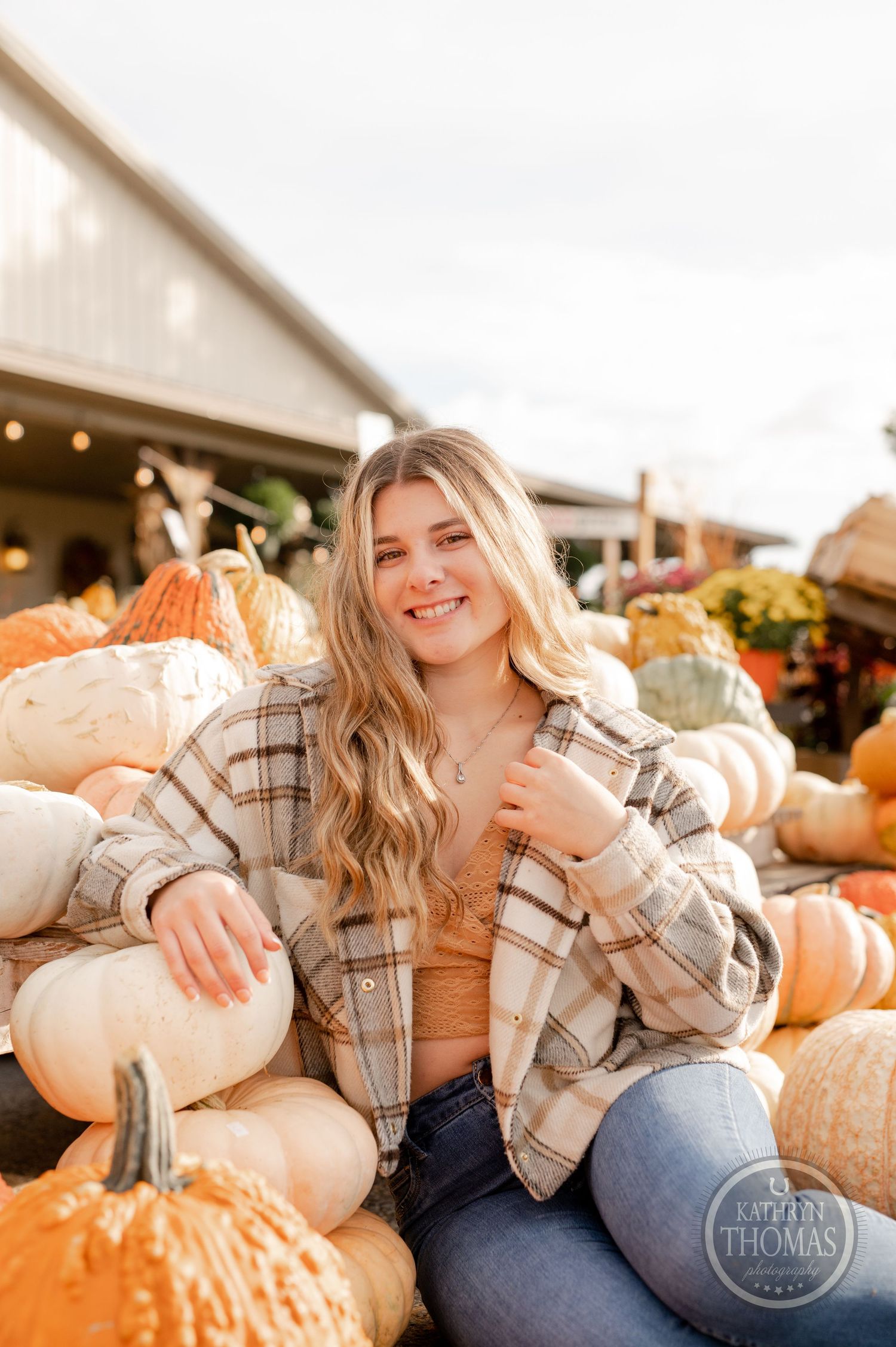 A casual autumn portrait surrounded by orange and white pumpkins at a rustic farm stand.