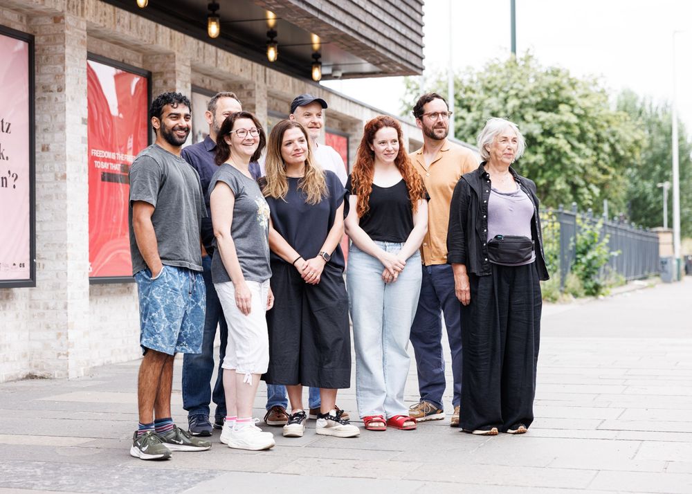 Group of seven people posing together outdoors on a sunny day near a building with red signage.