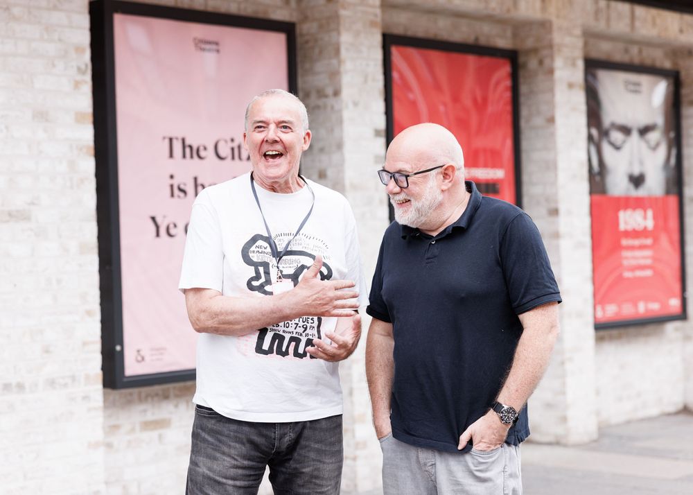 Two men laughing together outside a venue with promotional posters on the wall behind them.
