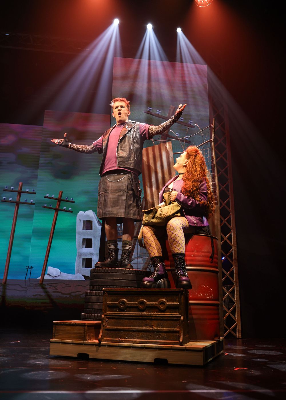 Two performers act out an emotional scene on a multi-level stage set with dramatic lighting and telephone poles in background.