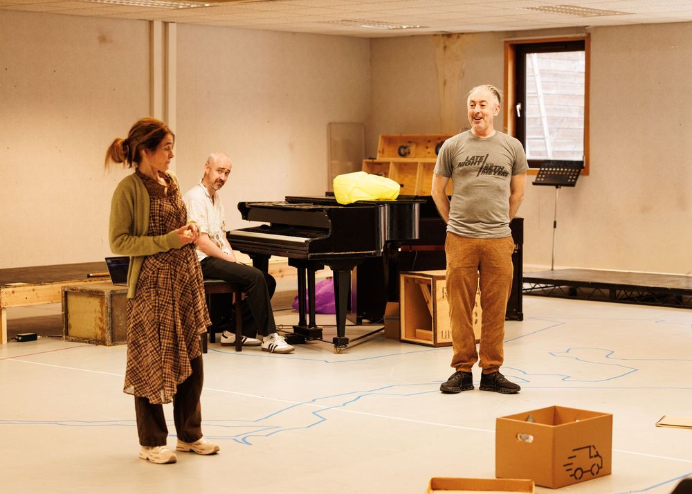 Three people in a rehearsal room with a grand piano, two standing and one seated, in a spacious studio space.