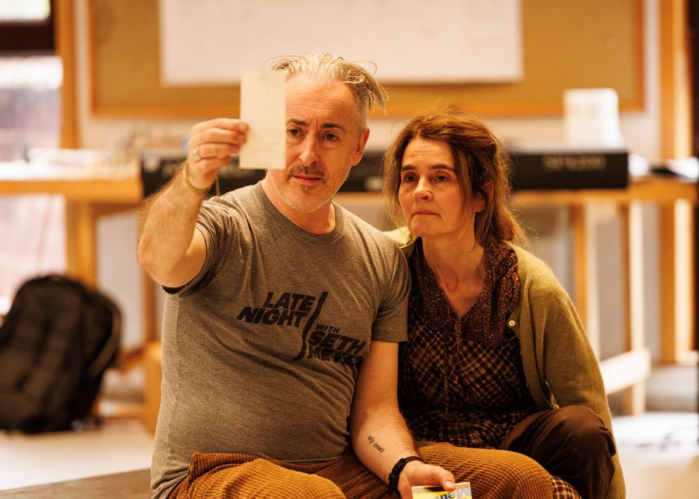 A man in a Late Night t-shirt holds up a paper while sitting beside a woman with braided hair indoors.