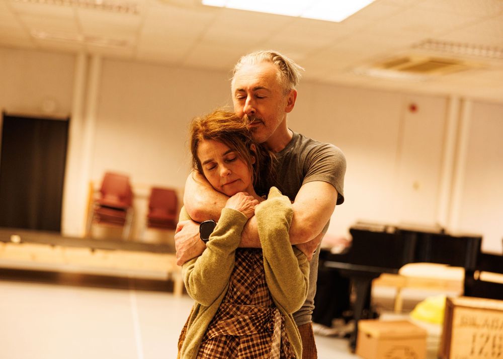Two actors rehearse an emotional embrace scene in a rehearsal room, with a man hugging a woman from behind.