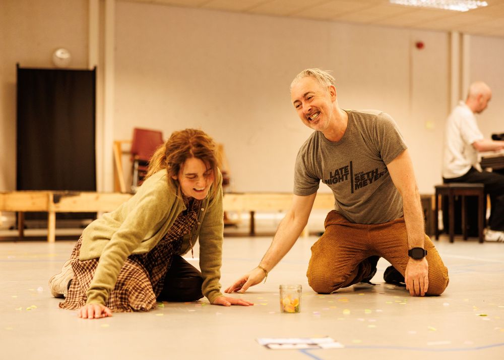 Two actors rehearse on a studio floor, smiling and gesturing during a theater rehearsal session.