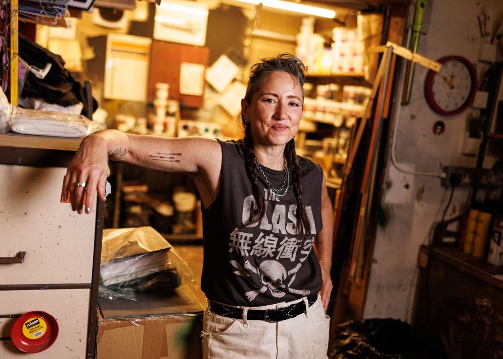Person in The Clash tank top leans against shelving in a cluttered workshop, showcasing tattoos and casual style.