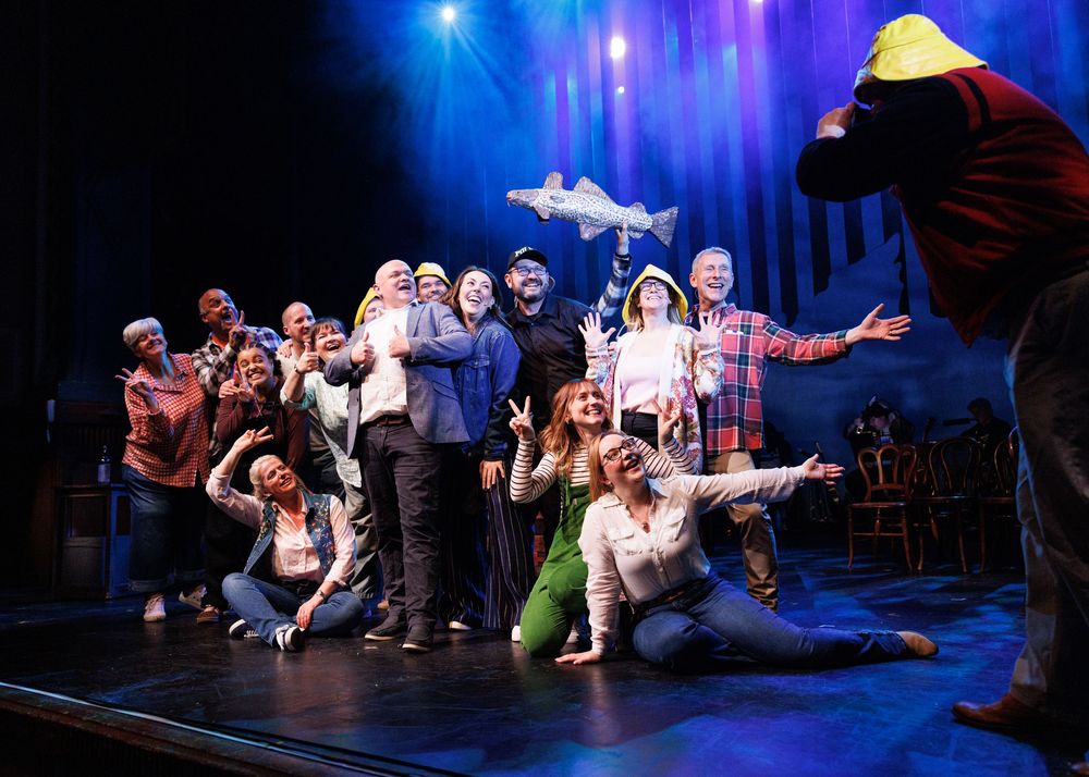 A diverse theater cast poses joyfully on stage during a group photo, with one actor tossing a prop shark in the air.