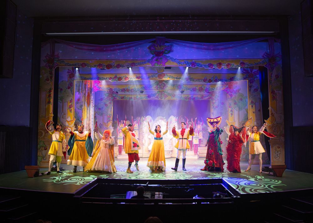 Musical theater performers in period costumes stand on stage with dramatic blue lighting.