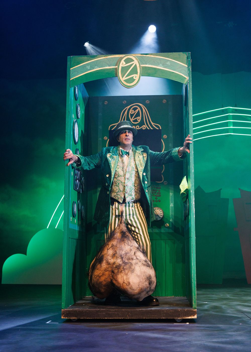 Stage performer in striped costume stands dramatically in a green-lit doorway prop with illuminated design elements.