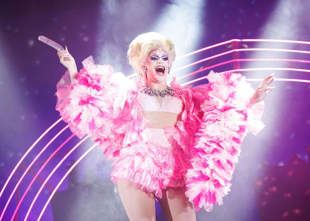 Performer in a glittering costume with pink feathers dances on stage under dramatic purple lighting effects.