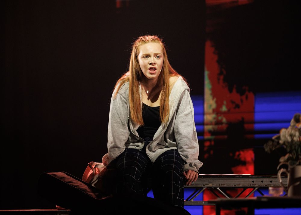 Young actress performing on stage in a gray hoodie and plaid pants under dramatic red and blue lighting.