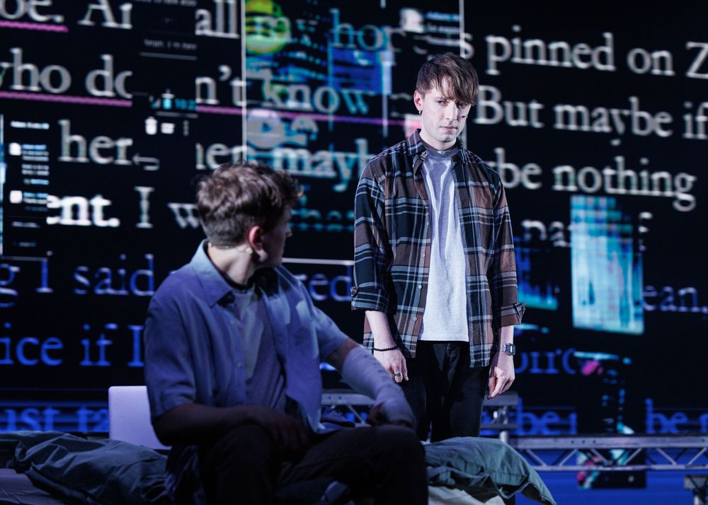 Two actors perform on stage with projected text backdrop in a dramatic theater production scene.