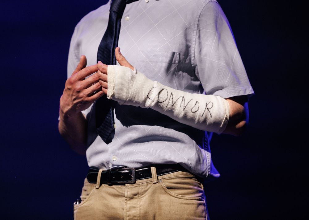 Person in gray shirt and khakis with bandaged arm reading CONNOR, holding a tie on stage.