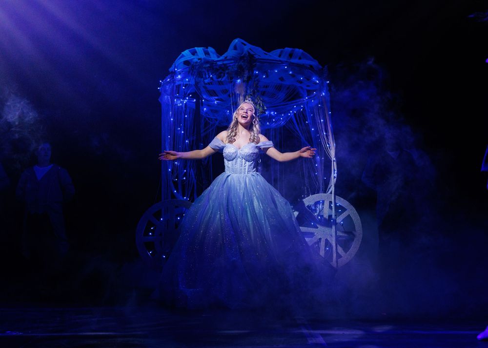 A performer in a sparkly blue gown twirls on stage during a theatrical production with dramatic blue lighting.