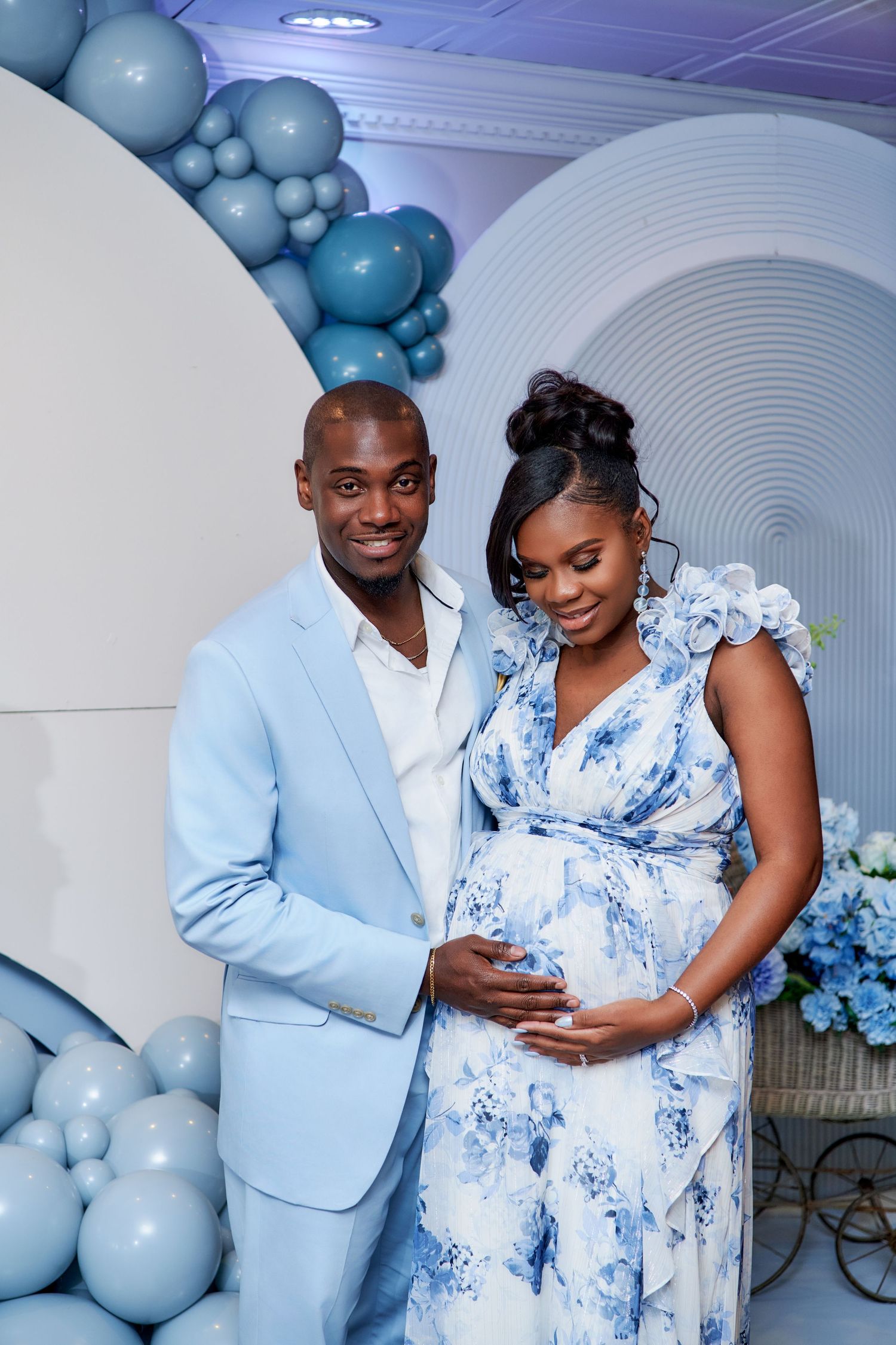 Expecting couple poses at baby shower with blue balloons and floral decorations against white backdrop.