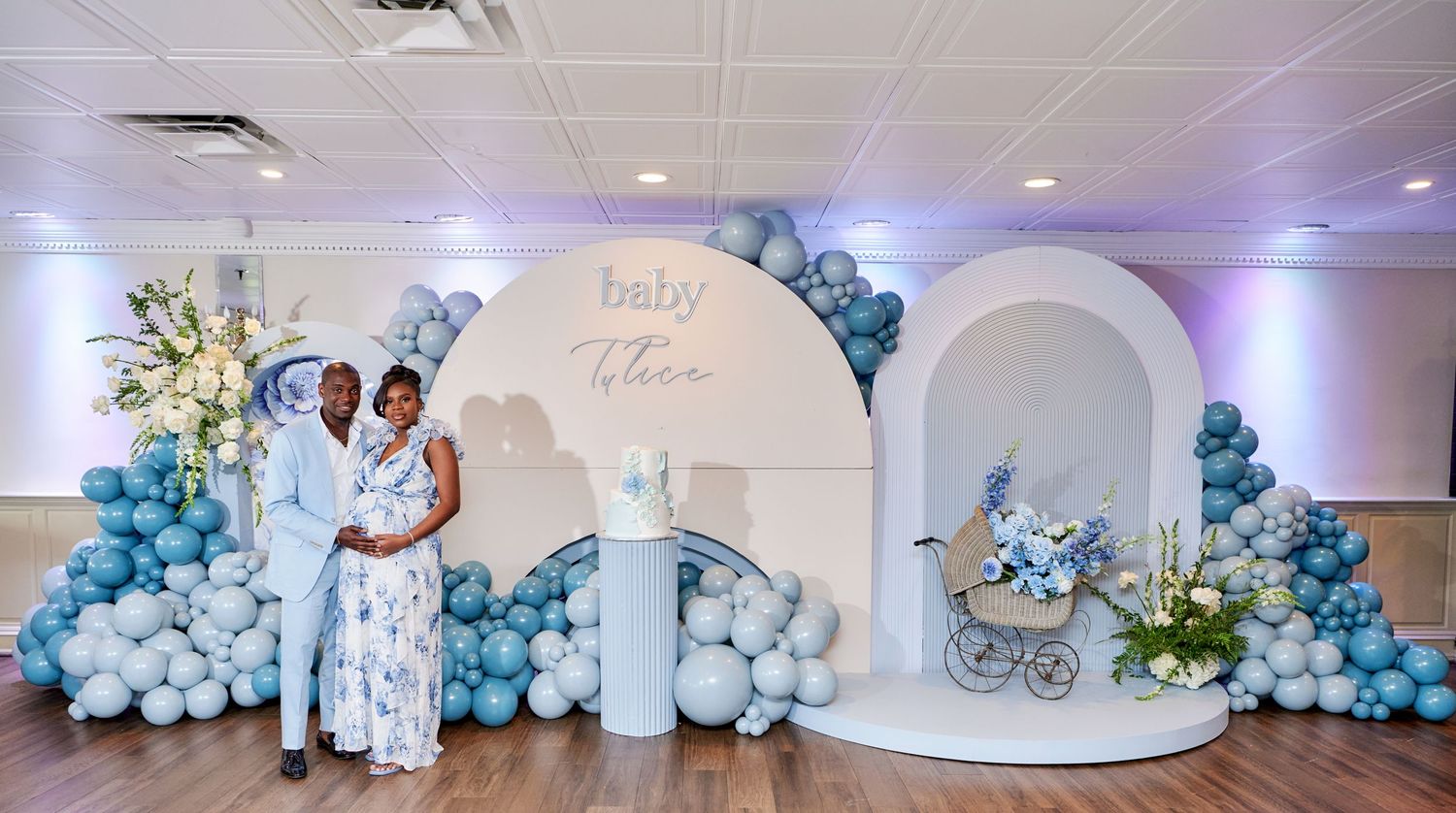 Elegant baby shower backdrop with white arches, blue balloon garland and floral decorations at a party venue.