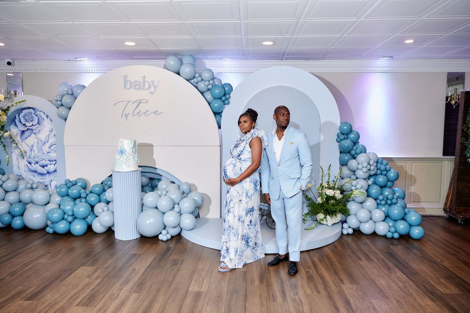 Couple poses in front of elegant white arch with blue balloon decorations at indoor celebration event.