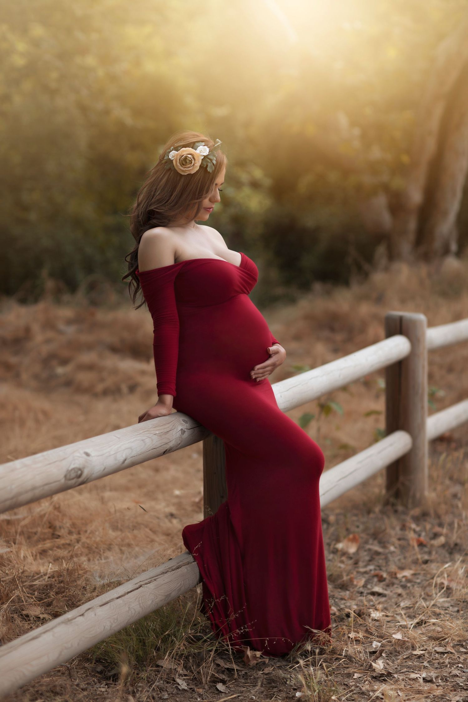Romantic maternity photo of woman in red dress leaning against a wooden fence at sunset.