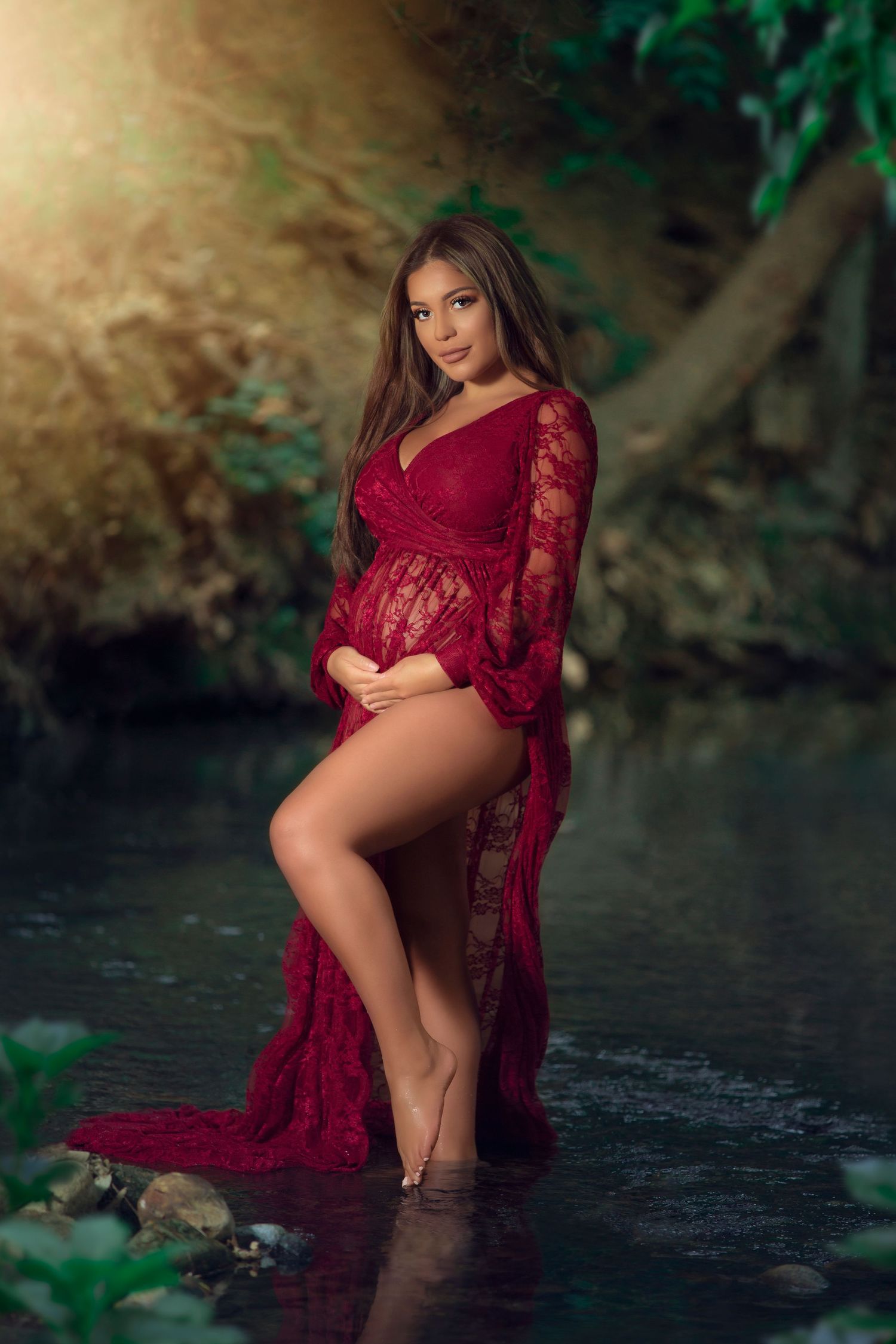 Maternity photoshoot in nature featuring burgundy lace dress against a scenic waterside background.