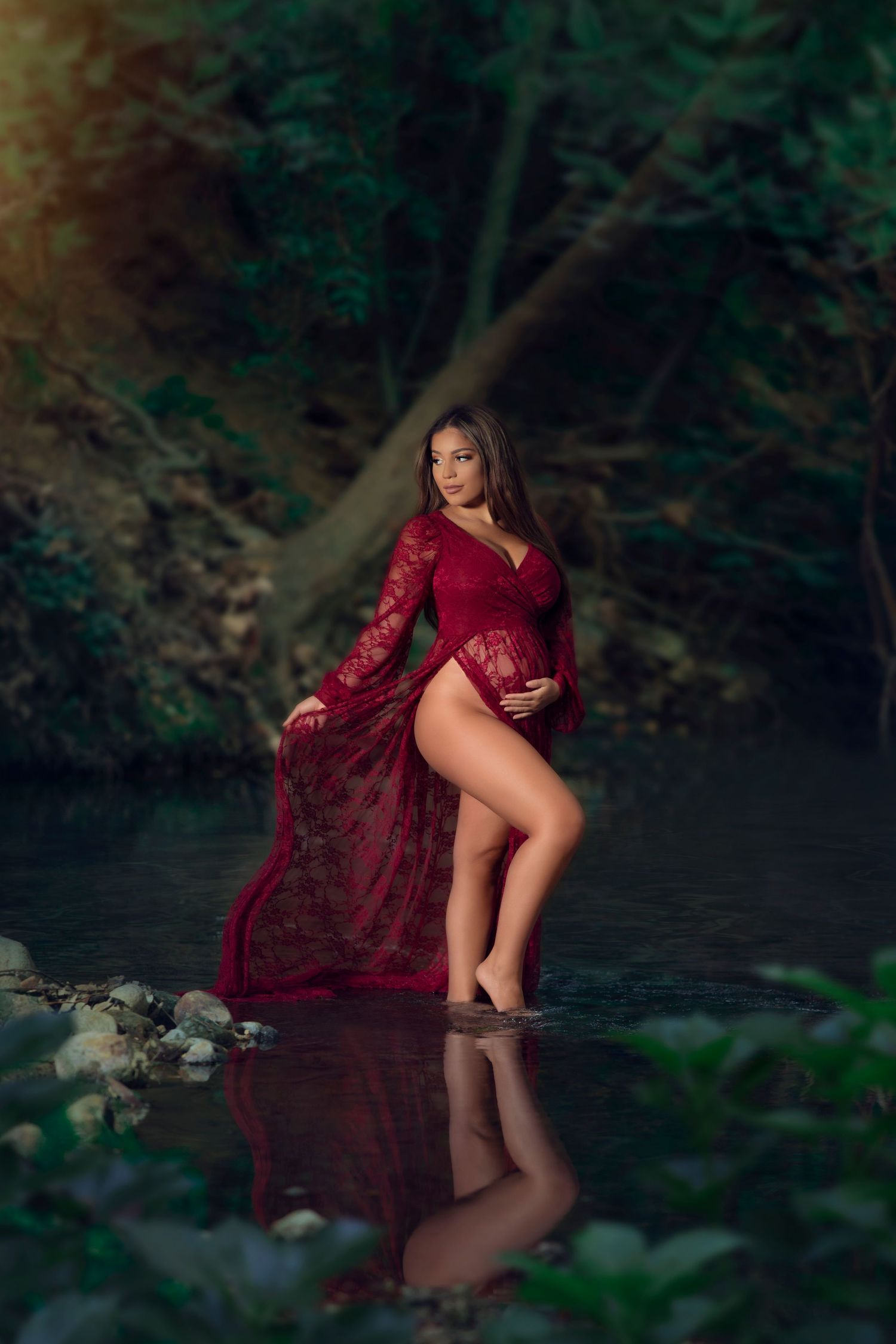A dramatic maternity portrait in flowing red dress at waters edge in forest setting.