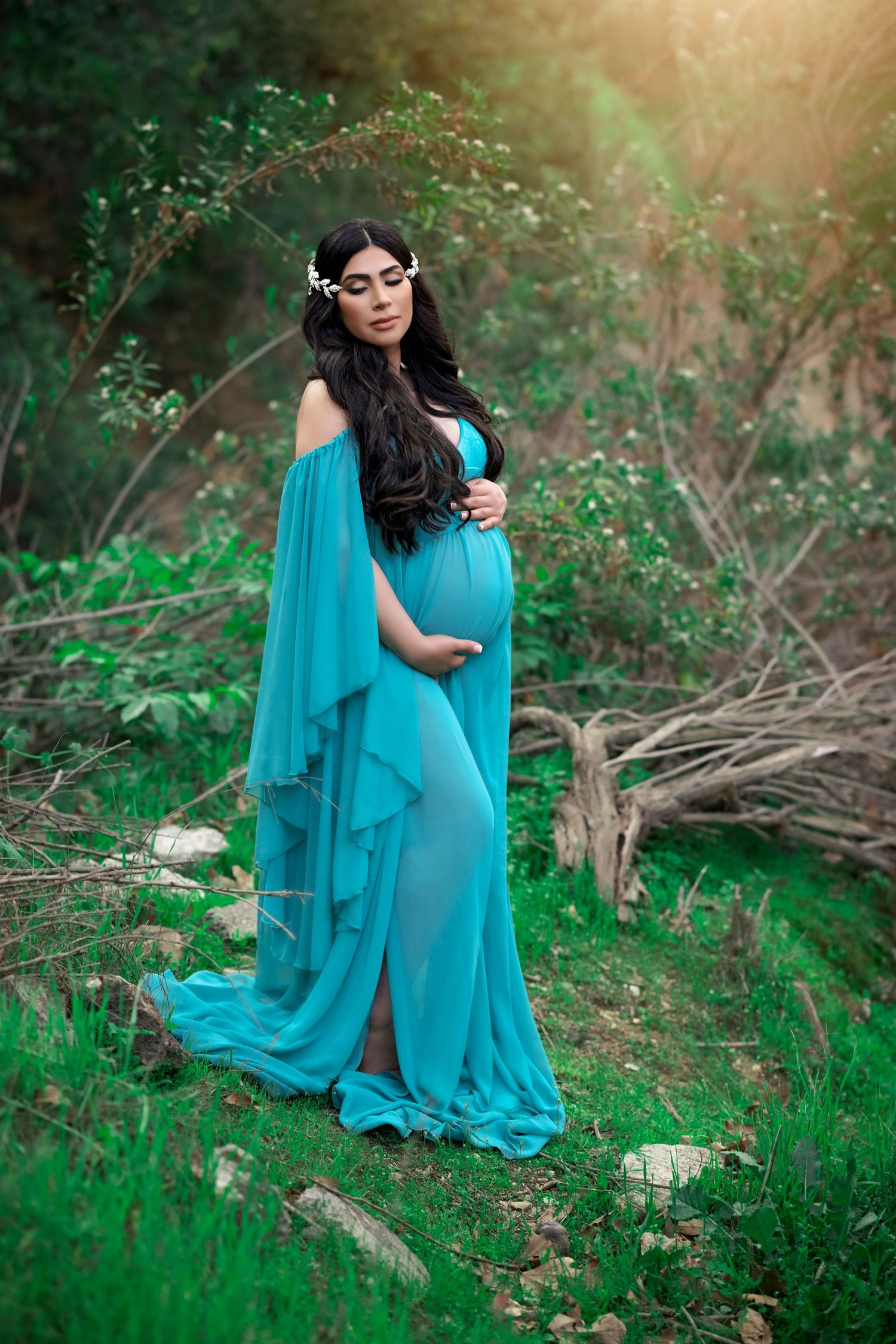 A dreamy maternity portrait in a flowing turquoise dress amid lush greenery.