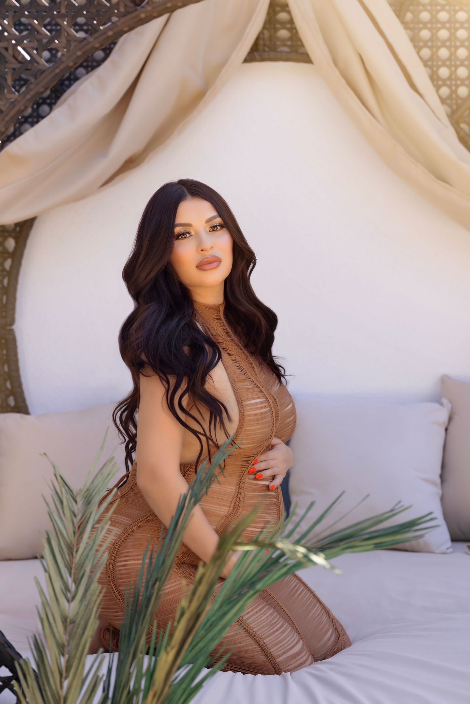 Boho maternity portrait with tan fitted dress surrounded by tropical leaves and white drapes.