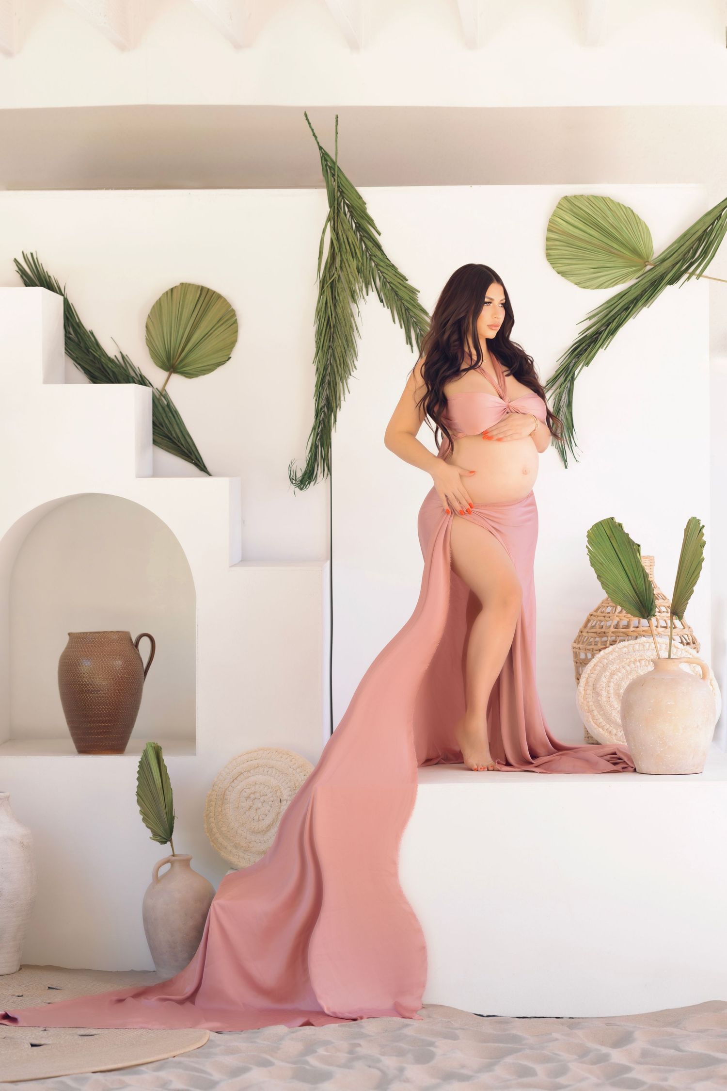 Bohemian maternity photo with pink flowing dress against Mediterranean-style white walls and palm decor.