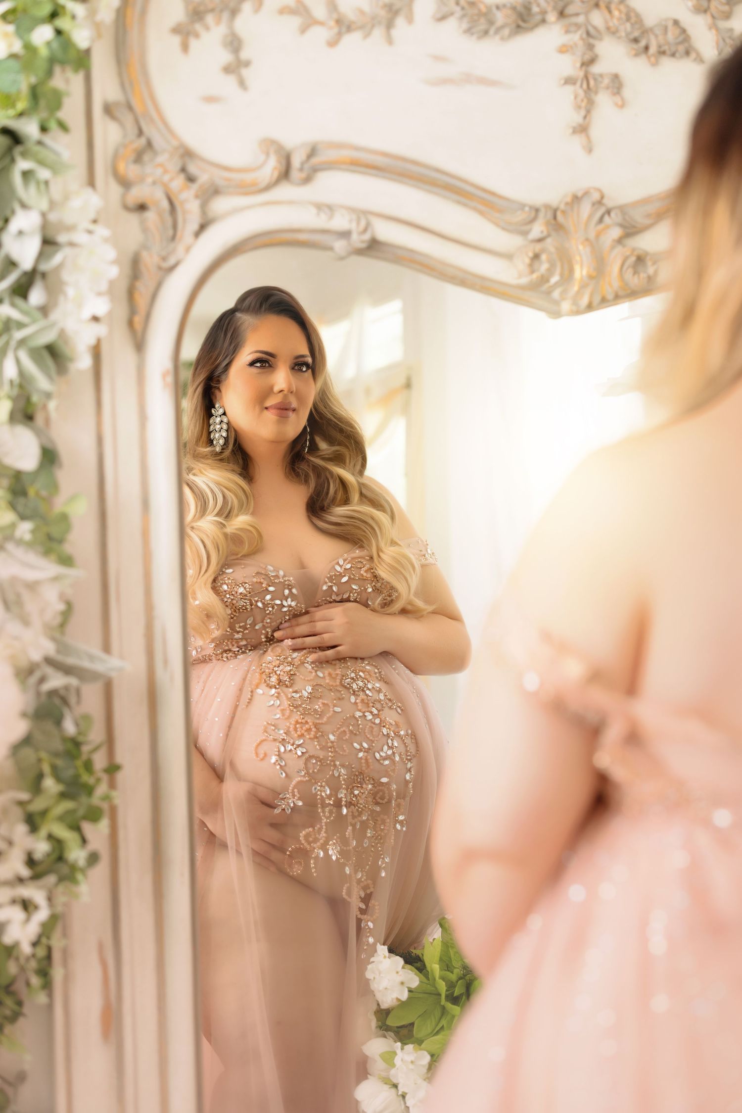 Elegant maternity portrait reflected in an ornate mirror surrounded by floral decorations.