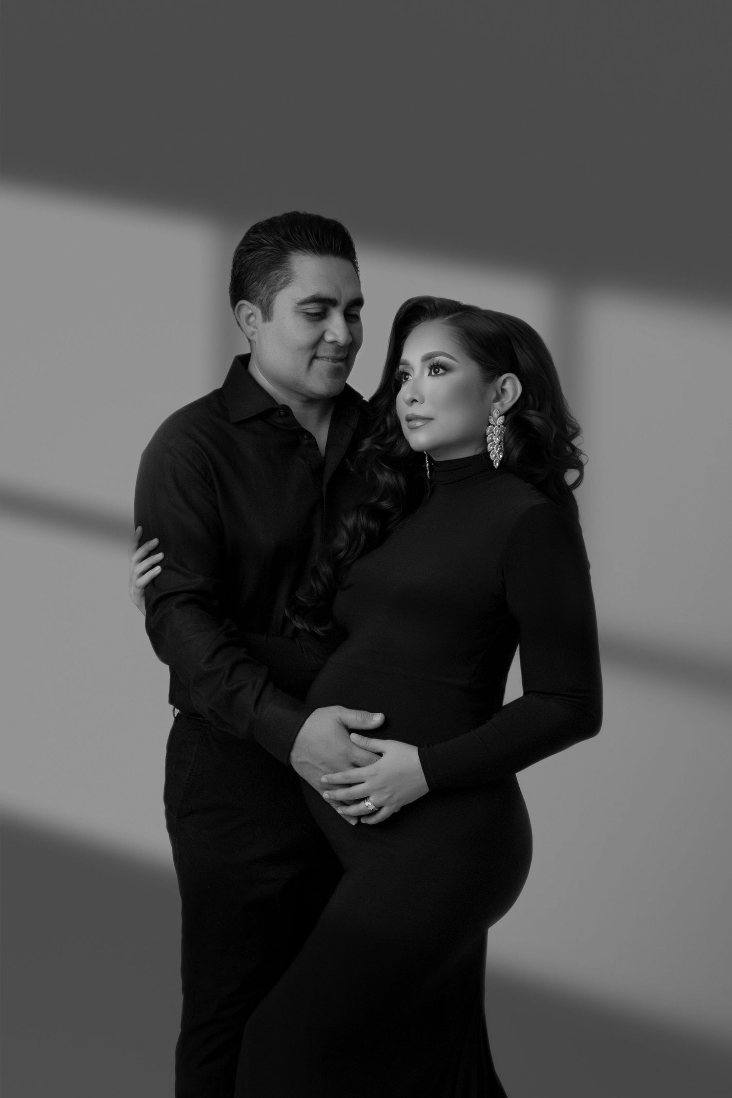 Black and white maternity portrait of couple in dark attire with dramatic lighting.