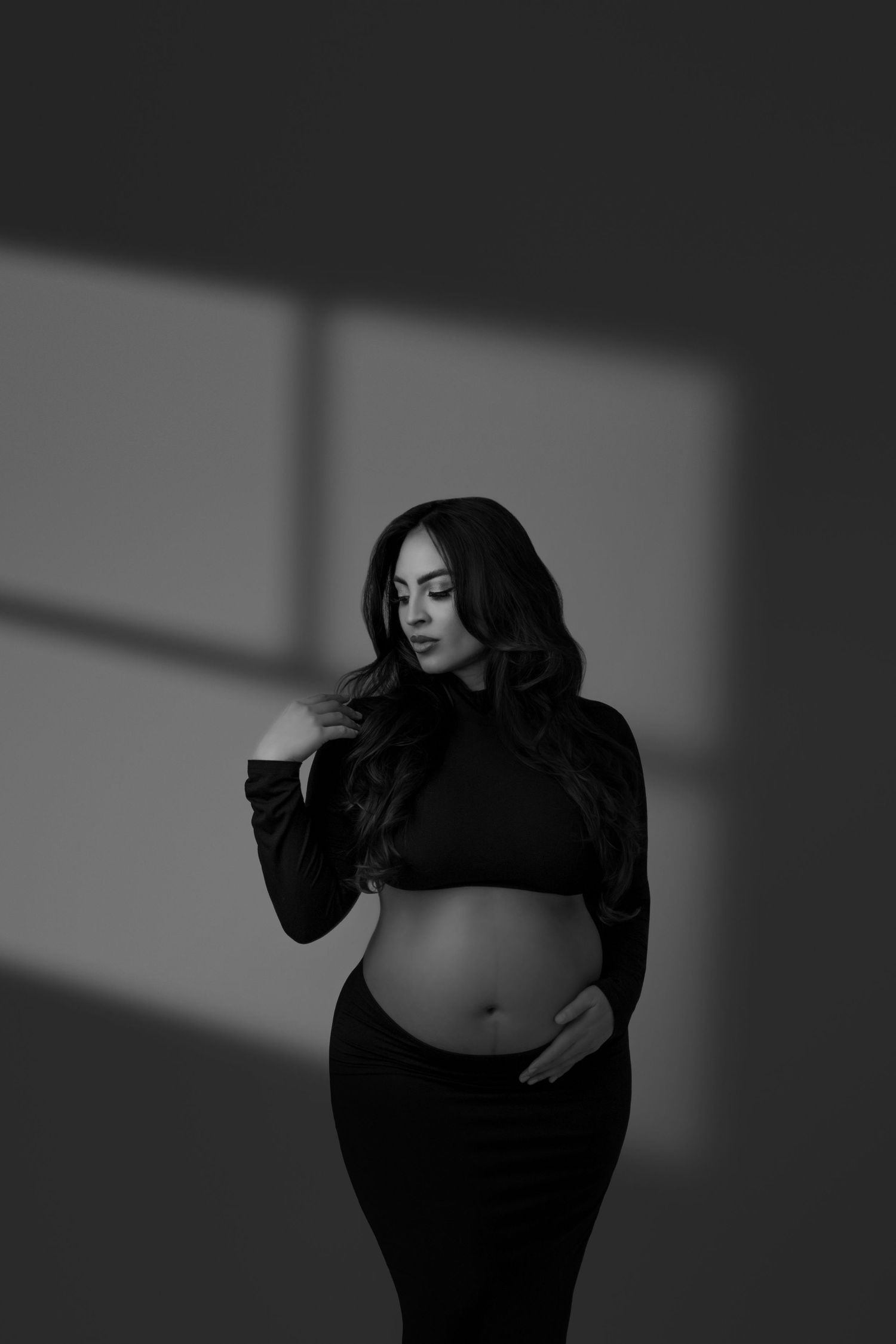 Artistic black and white maternity photo showing an expectant mother in a cropped black top against dramatic shadows.