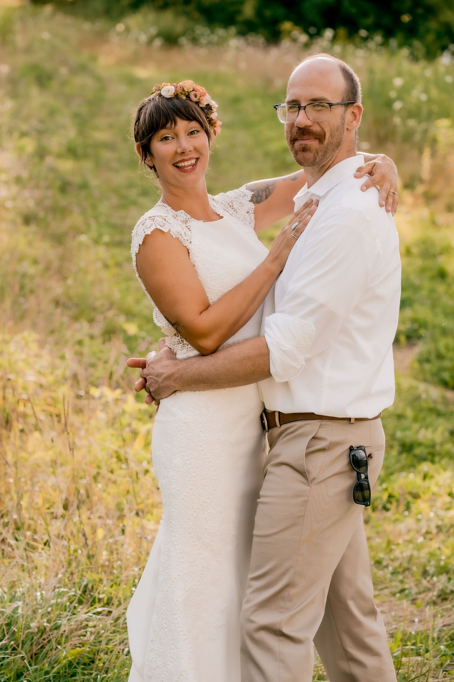 A couple in wedding attire embraces outdoors in a sunlit field with natural greenery in the background.