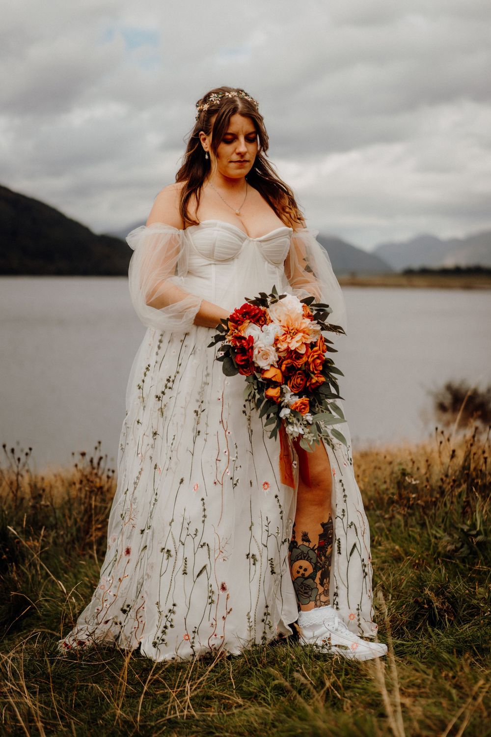 A bride in a flowing white dress holds an autumn-colored bouquet against a moody lakeside landscape with mountains.