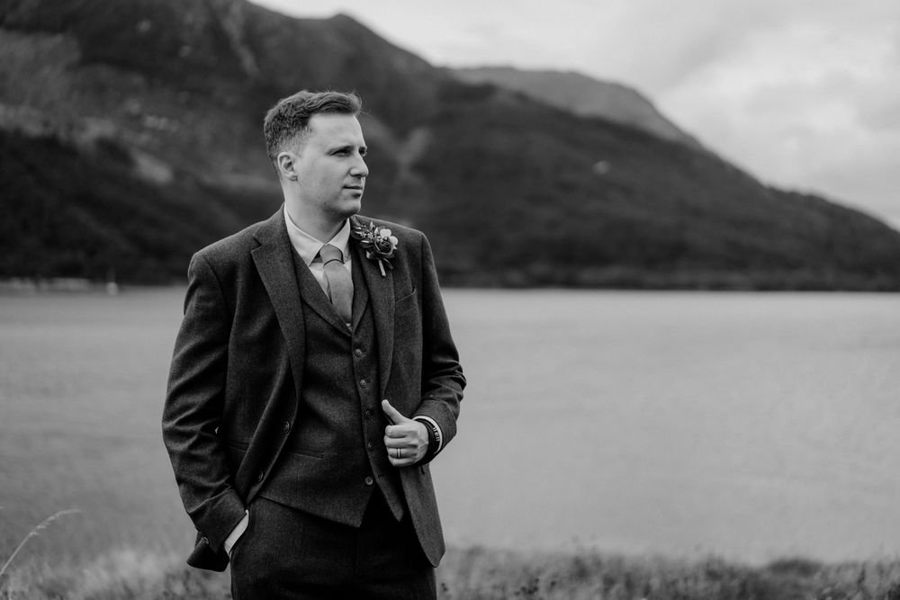 A well-dressed person in a dark suit stands thoughtfully against a scenic mountain lake backdrop.