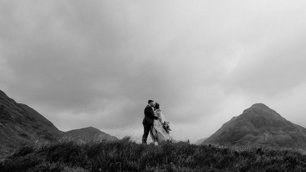 A silhouetted couple stands on a hilltop between two mountain peaks under dramatic cloudy skies.