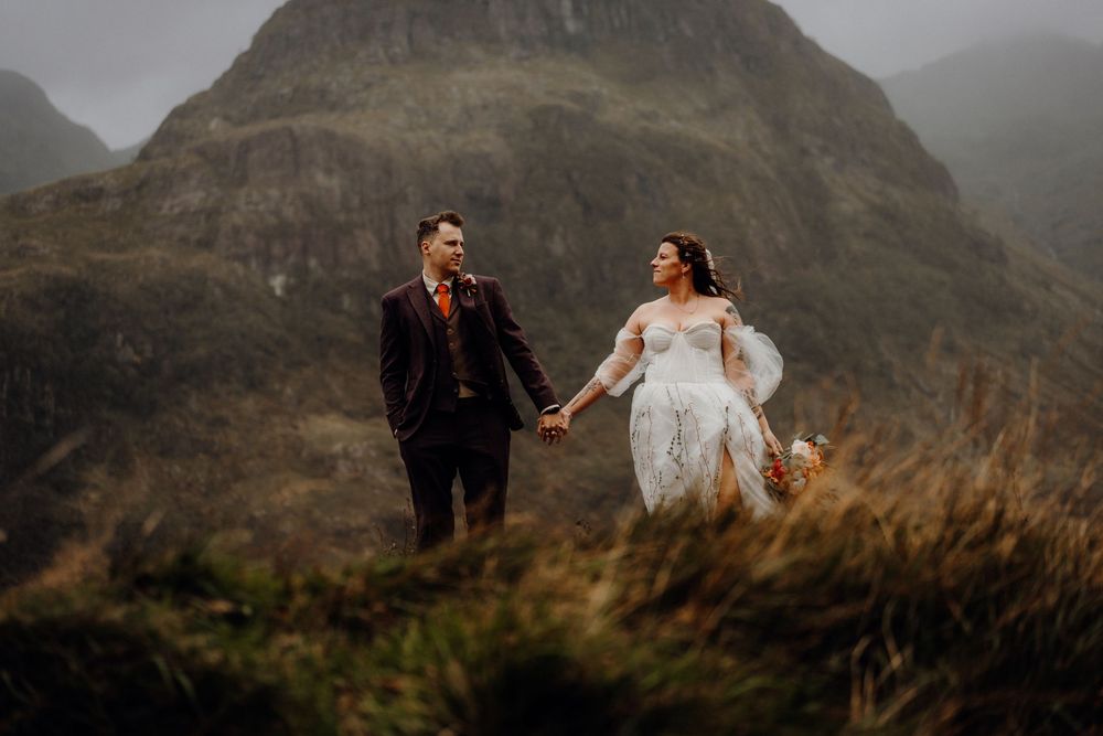 A couple poses together in dramatic mountainous terrain as wind blows through a flowing white dress.