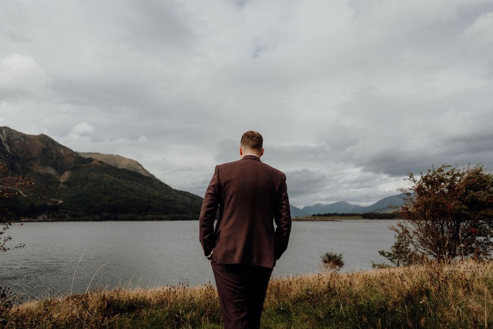 A person in brown jacket stands at scenic mountain viewpoint overlooking misty lake and rugged peaks on cloudy day.
