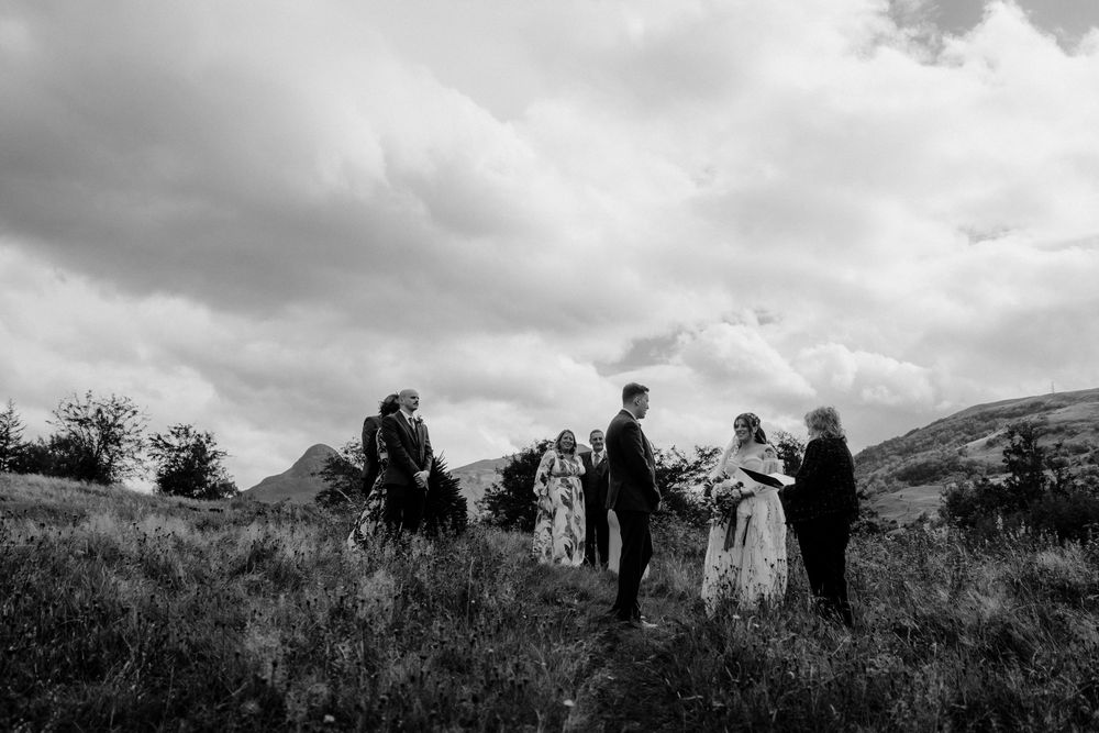 A small wedding ceremony takes place on a hilltop meadow with dramatic mountains and moody clouds in the background.