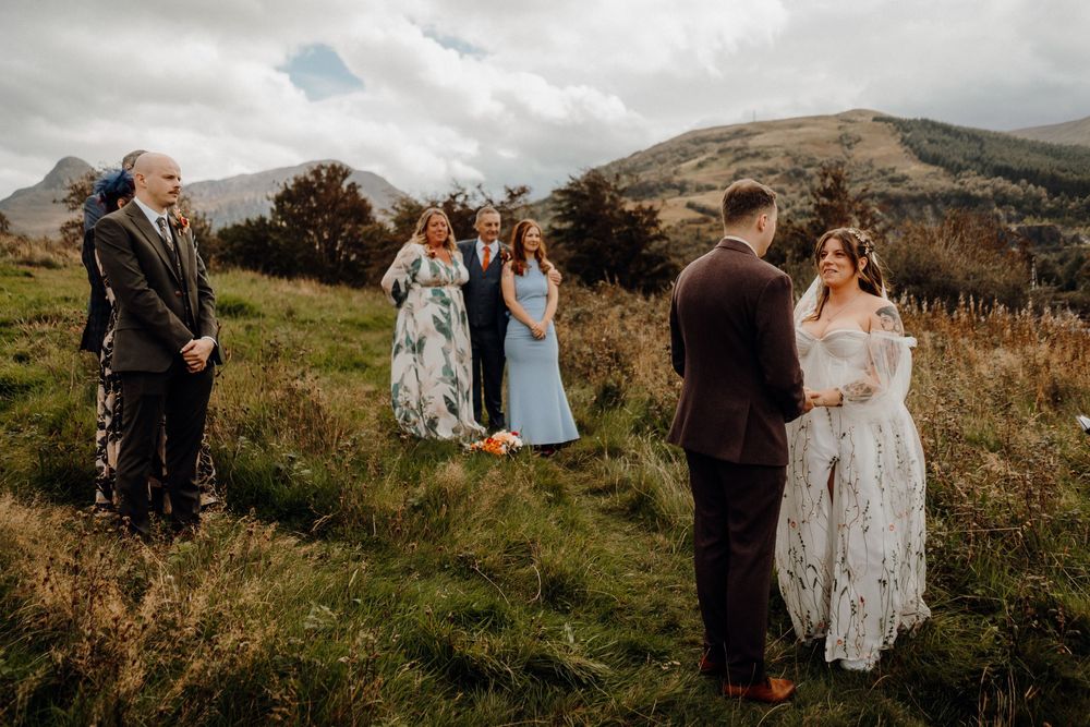 A couple shares an intimate moment on a grassy hillside overlooking a scenic mountain landscape during their wedding.