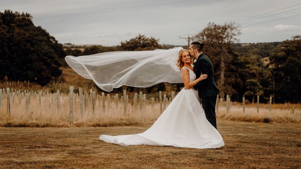 A flowing white wedding veil billows in the wind across a rustic outdoor field setting at sunset.
