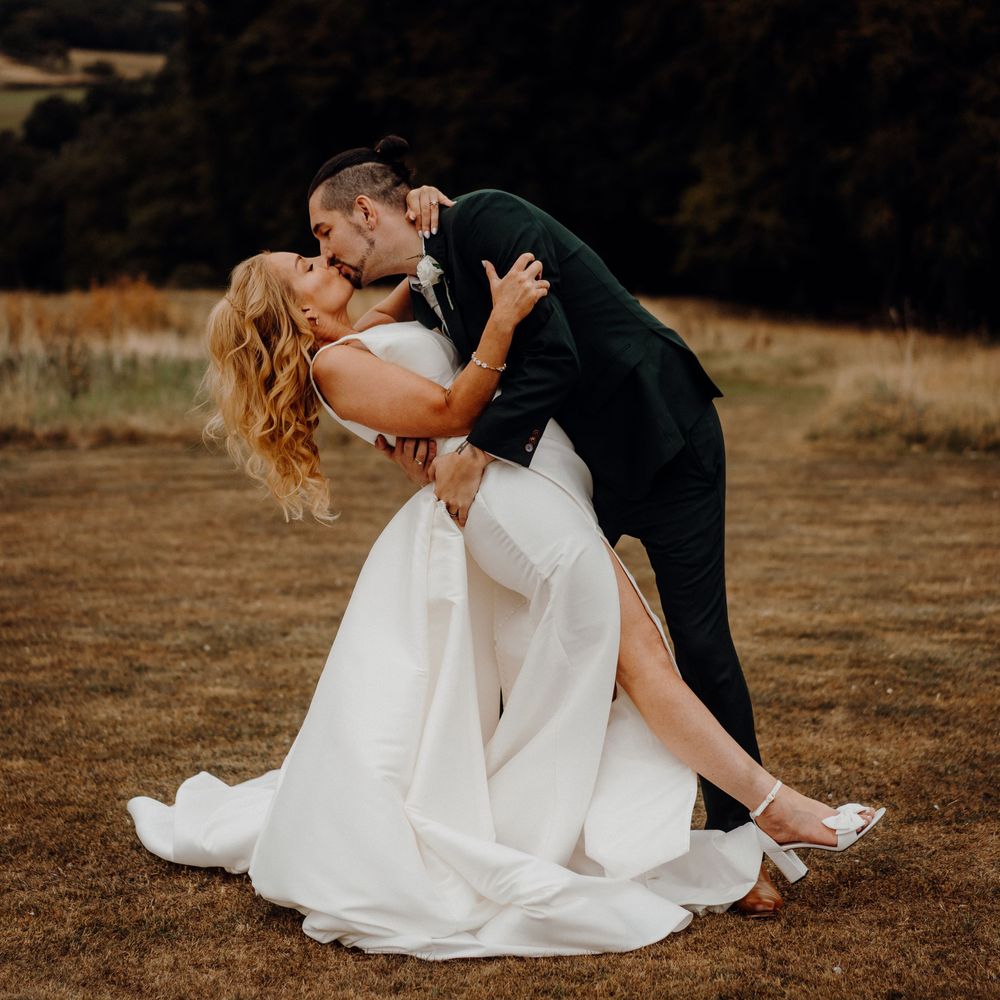 Romantic couple in wedding attire share intimate dips and kisses in a rustic outdoor setting at sunset.
