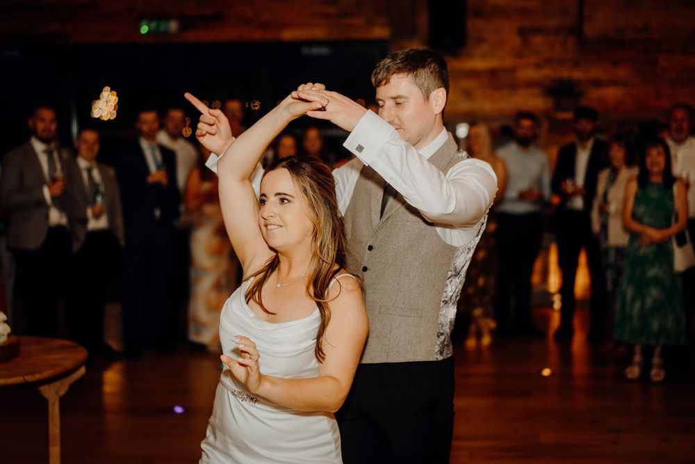 A couple dances together at an indoor wedding reception with warm lighting and wooden beams overhead.
