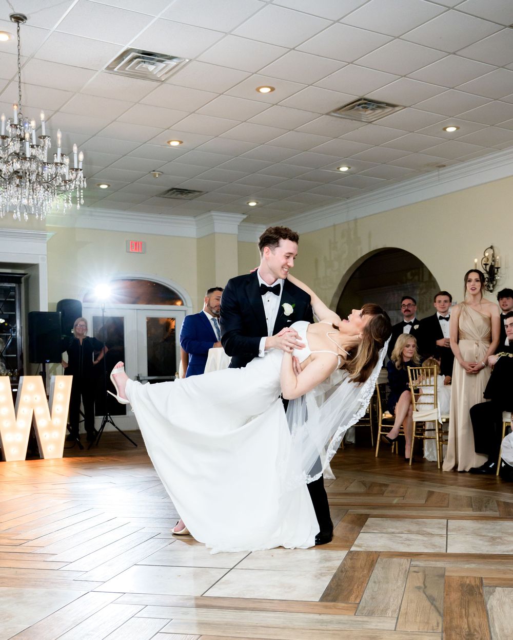 A couple performs a romantic dip during their first dance at an elegant wedding reception with a chandelier overhead.