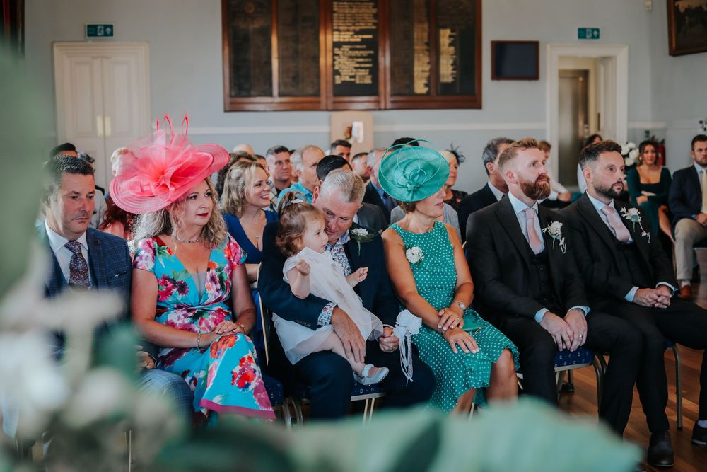 Wedding guests in colorful attire seated in rows during a ceremony with vibrant floral hats.