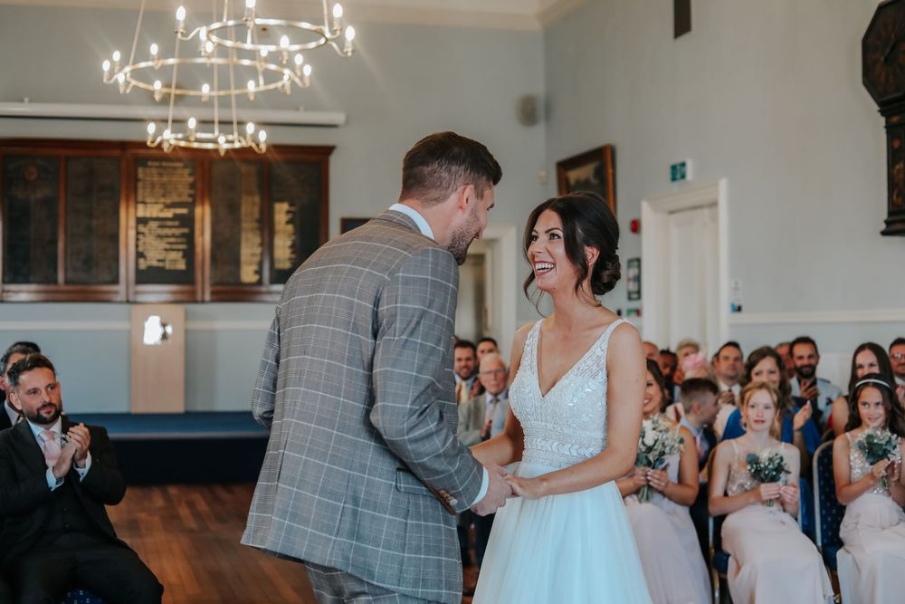 Wedding ceremony in a elegant hall with chandelier as couple exchanges vows in front of seated guests.