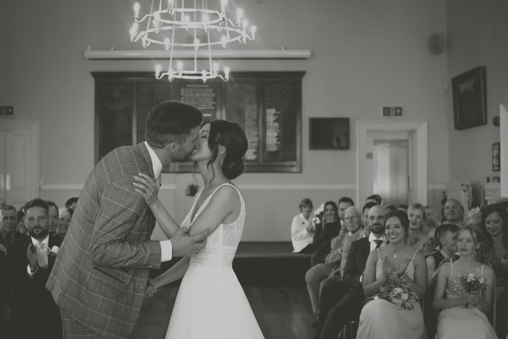 Black and white photo of tender moment during wedding ceremony with guests watching in background.