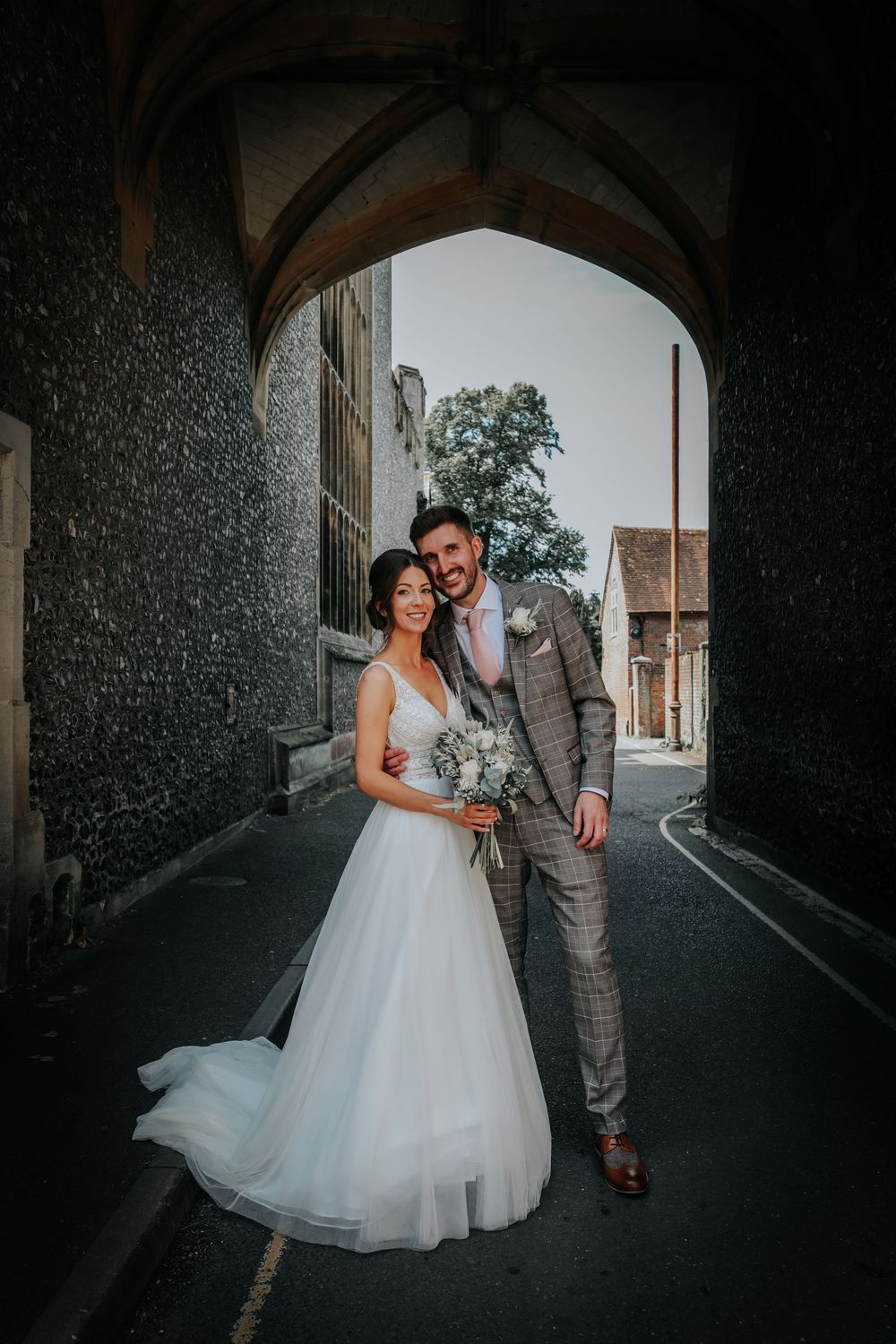 Newlyweds pose under stone archway in elegant wedding attire with natural lighting.