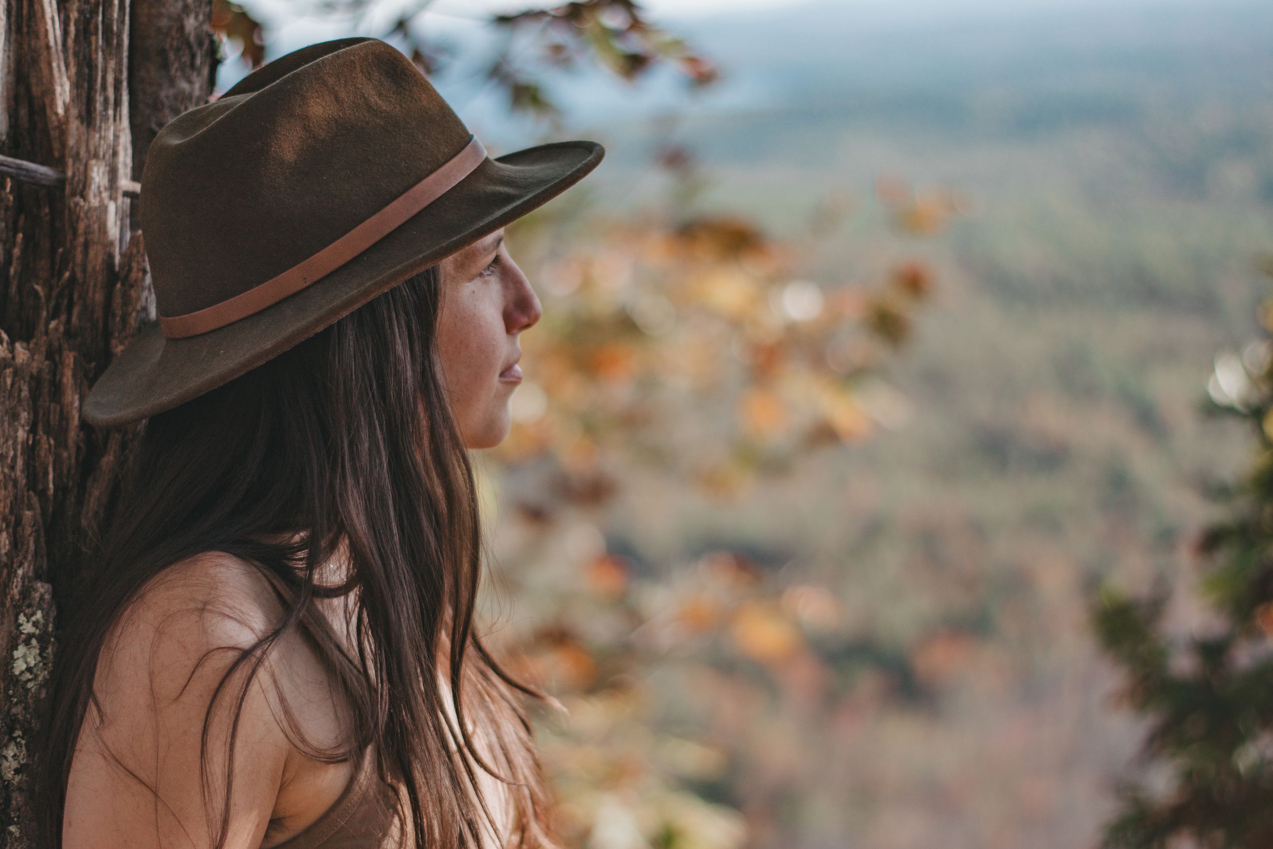 A young woman in a wide-brim hat gazes at an autumn forest landscape from a scenic overlook.