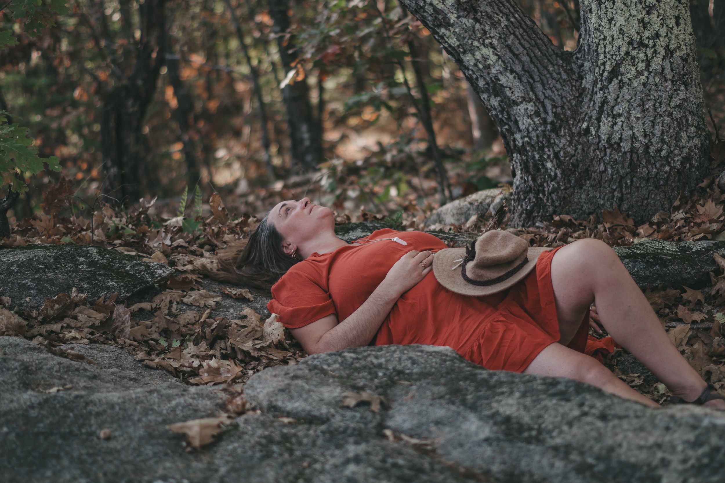 Woman in red dress reclining on rocks in autumn forest with hat on chest.